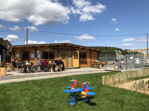 Im Hintergrund sieht man eine große eingeschossige Holzhütte . In der Mitte ist eine Doppeltür. Darüber hangt ein Holzschild mit der Aufschrift "Bielenhof Alm". Rechts und links der Tür sind Flügelfenster mit Blumenkästen. Vor der Hütte gibt es eine gepflasterte Terrasse mit einigen Tische und Stühlen.Zwei der Tische sind mit einer Männergrppe belegt.Im Vordergrund steht auf einer Rasenfläche eine Federwippe n den Farben blau, gelb und rot.i