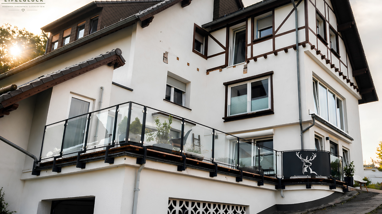 A modern house with a large balcony and glass railing. The facade is brightly painted and features traditional roof details.