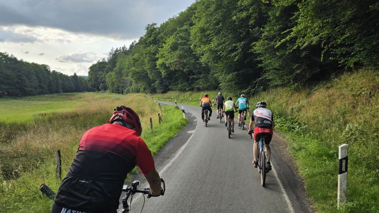 Gruppe von Radfahrern auf einer Landstraße in der Eifel, umgeben von grünen Wiesen und Bäumen.