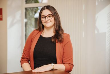 Eine Frau steht hinter einem Tresen in einem hellen Büroraum und lächelt in die Kamera. Hinter ihr einige Fenster.