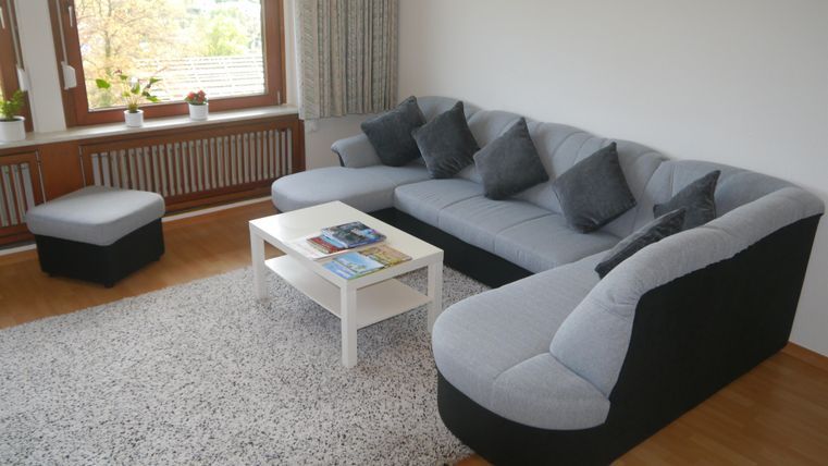 Ein gemütliches Wohnzimmer mit einem großen, grauen Sofa und einem weißen Tisch. Auf dem Teppich liegen einige Bücher, und es gibt Fenster mit Pflanzen im Hintergrund.