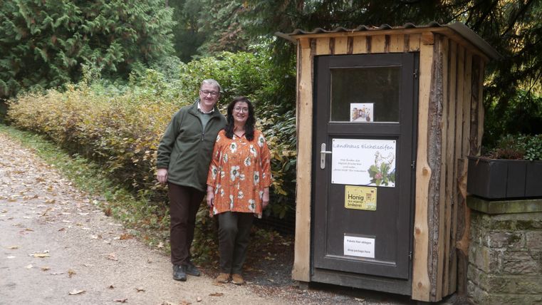 Ein Mann und eine Frau stehen neben einem kleinen Holzhaus mit Aufschrift Landhaus Eichelseifen.