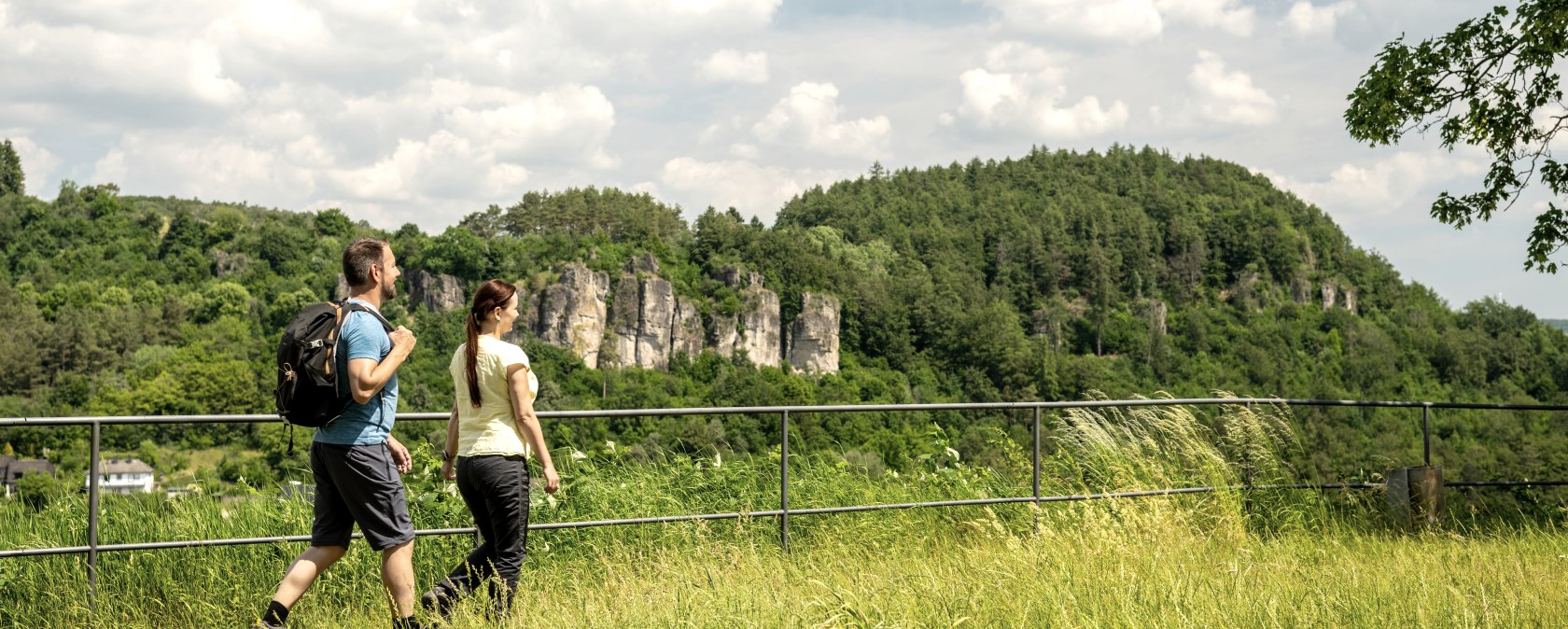 Zwei Wanderer auf einer Wiese neben einem Zaun. Im Hintergund sind hohe Felsen umgeben von B&auml;umen zu sehen.