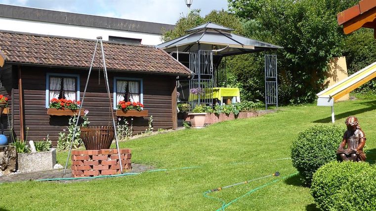 Ein schöner Garten mit einem historischen Holzhaus und bunten Blumenkästen. Im Hintergrund steht ein Pavillon und eine Rutsche für Kinder.