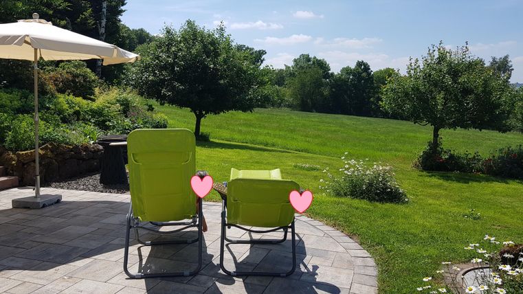 Ein schöner Garten mit zwei grünen Liegen und einem Sonnenschirm. Im Hintergrund sind Bäume und eine weitläufige Wiese zu sehen.
