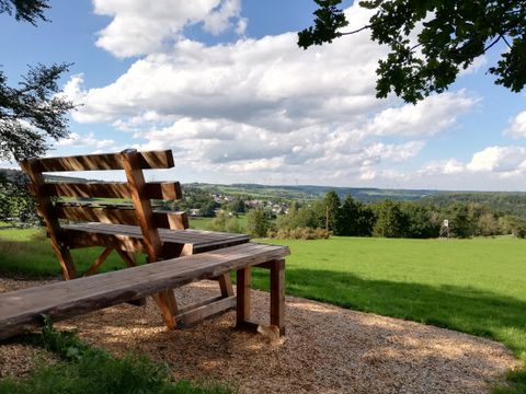 Eine große Holzbank steht auf einem Hügel mit Blick auf eine grüne Landschaft und einen bewölkten Himmel.