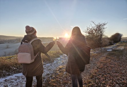 Two people stand on a hiking trail covered with a little snow and clink two wine glasses together. In the background, the sun sets behind fields.