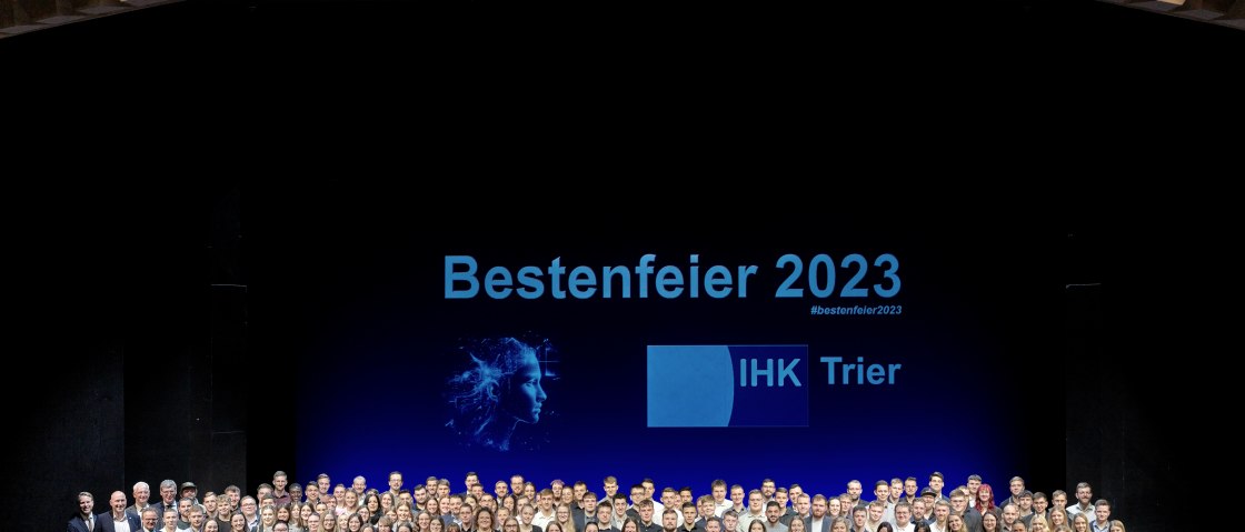Zahlreiche Personen stehen dicht beieinander auf einer B&uuml;hne und halten Auszeichnungen in den H&auml;nden. Hinter Ihnen der Schriftzug "Bestenfeier 2023 IHK Trier"
