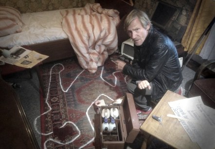 Ein Mann kniet in einem Schlafzimmer auf dem Boden. Das Zimmer ist unordentlich verwüstet und vor im ist der Umreiss einer Leiche mit kreide auf den Teppich gemalt.