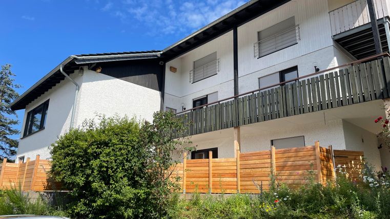 Un immeuble résidentiel moderne avec un balcon et un environnement verdoyant. Le ciel est clair et bleu.