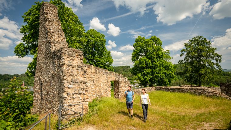 Zwei Personen spazieren an den Ruinen der Löwenburg in Gerolstein vorbei, umgeben von grünen Bäumen und blauem Himmel.