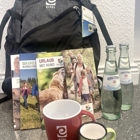 Een Eifel-rugzak met verschillende toeristische brochures, twee Eifel-bekers en twee glazen flessen Gerolsteiner Sprudel.