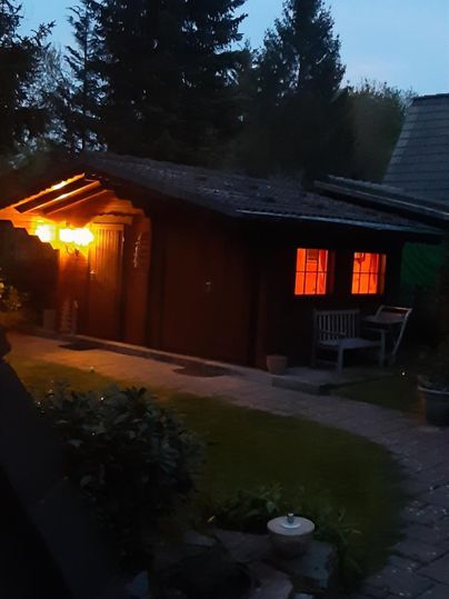 Ein gemütliches Holzhaus bei Dämmerung, beleuchtet von warmem Licht. Der Garten ist mit Pflanzen und Steinen dekoriert.