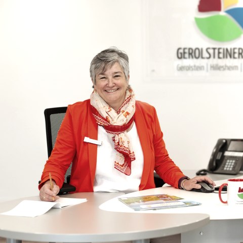 A woman sits at a desk in a bright office with a computer, telephone and keyboard in front of her and smiles at the camera. Behind her are brochure racks from the tourist information office and the logo of Touristik GmbH Gerolsteiner Land.