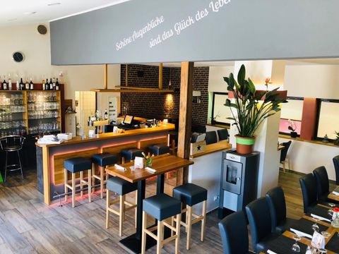 Modernes Restaurant mit Bar, Holztischen und Stühlen. An der Wand steht "Schöne Augenblicke sind das Glück des Lebens". Pflanzen und Gläser dekorieren den Raum.