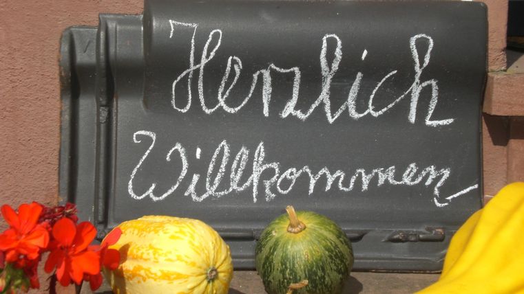 Ein Willkommensschild aus schwarzem Brett mit der Aufschrift „Herzlich Willkommen“. Daneben liegen bunte Gemüse und Blumen.
