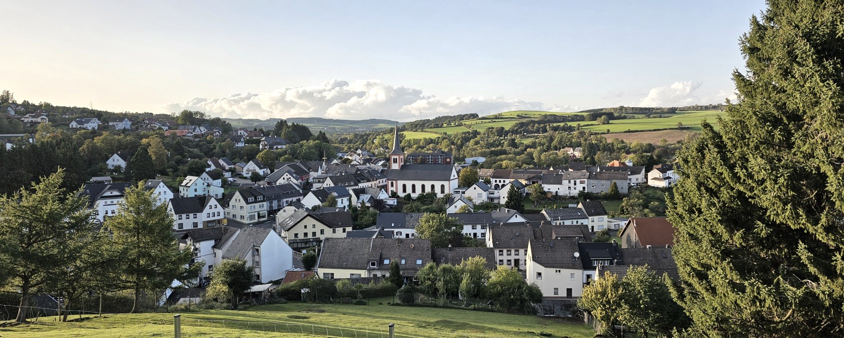 Panoramablick auf Stadtkyll mit Kirche im Zentrum, umgeben von grünen Feldern und Bäumen unter blauem Himmel., © Touristik GmbH Gerolsteiner Land, Leonie Post