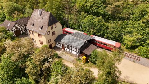 Un train rouge est stationné à côté d'un ancien bâtiment de gare, au milieu de beaucoup de verdure.