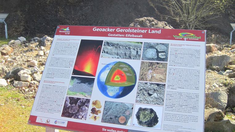 Infotafel im Geoacker Gerolsteiner Land über Eifelbasalt, mit Bildern und Texten zur Entstehung und Eigenschaften des Basalts. Hintergrund: Felsen und Vegetation.