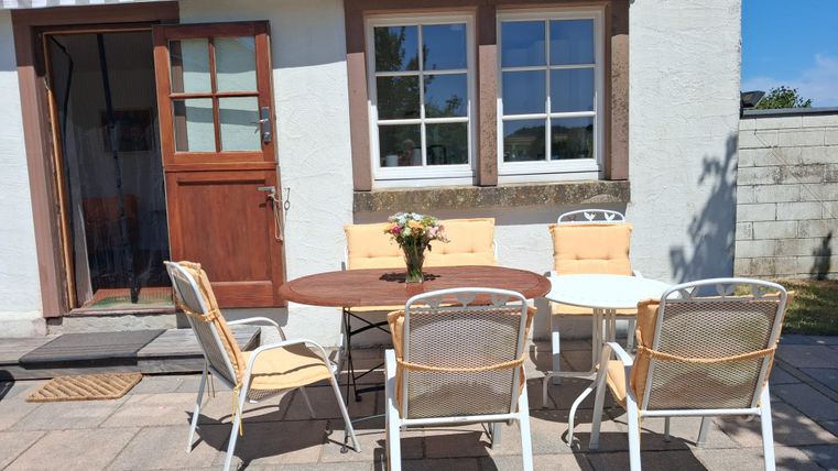 Ein gemütlicher Außenbereich mit einem Tisch und Stühlen. Die Sonne scheint und es gibt eine Blumenmitte auf dem Tisch.