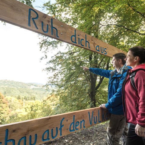 Zwei Wanderer stehen an einem Aussichtspunkt in einem Wald und zeigen in die Ferne. Sie stehen an einem Holzrahmen auf dem &quot;Ruh dich aus. Blick in den Vulkan&quot; steht.
