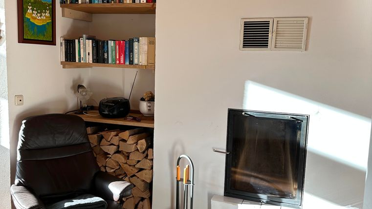 Ein gemütlicher Wohnraum mit einem schwarzen Sessel und einem Kamin. Im Hintergrund ist ein Bücherregal und eine Holzlege sichtbar.