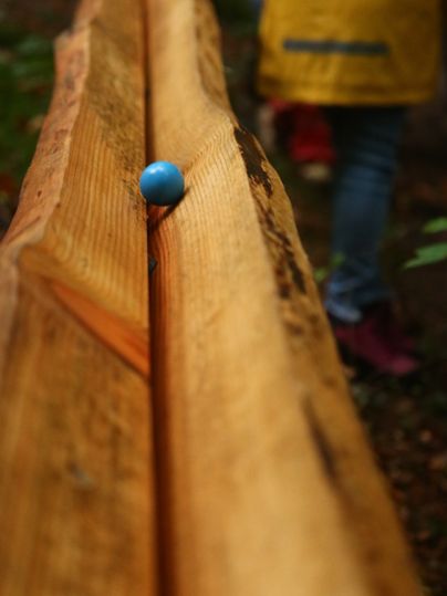 Zwei Holzbalken aneinander bilden ene Rille, auf der eine bunte Holzkugel rollt. Dicht daneben Laubbäume und ein Kind im Hintergrund.
