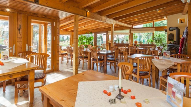 Gemütlicher Wintergarten mit Holzmöbeln, großen Fenstern und Pflanzen. Die Tische sind mit Tischdecken und Dekorationen geschmückt.