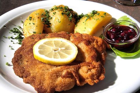 Ein Teller, auf dem ein großes paniertes Schnitzel mit einer Zitronenscheibe darauf liegt. Daneben drei Kartoffeln mit Kräutern und eine Schale mit Preiselbeeren darin.