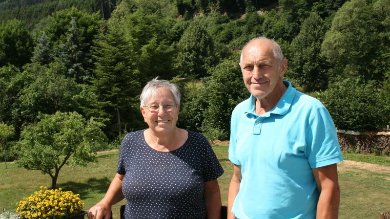 Ein älteres Paar steht in der Natur vor einem grünen Hintergrund. Die Sonne scheint, und die Stimmung wirkt freundlich und entspannt.