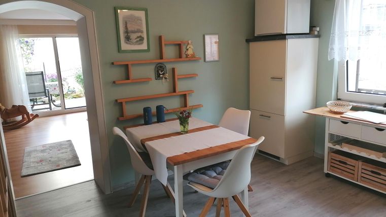 Eine helle Küche mit einem Tisch, Stühlen und einem Kühlschrank. Die Wände sind in einem sanften Grün gehalten und es gibt Holzregale für Dekoration.