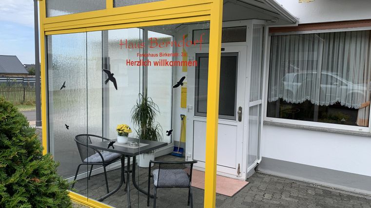 Ein einladender Eingangsbereich mit Glaswänden und einem kleinen Tisch mit Stühlen. Auf dem Tisch steht eine gelbe Blume und ein Willkommen-Schild ist sichtbar.