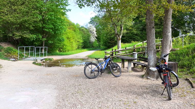 Zwei Fahrräder stehen auf einem Schotterweg neben Bäumen und einer Bank. Ein kleiner Bach fließt über den Weg. Umgeben von grünen Bäumen.