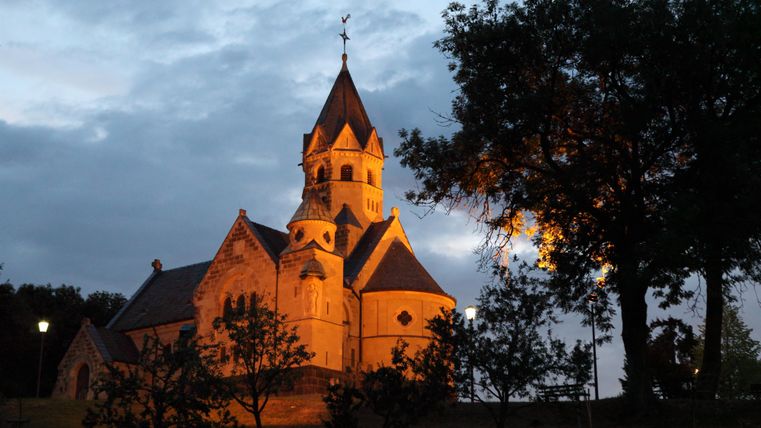 Die Erlöserkapelle Mirbach bei Dämmerung, beleuchtet von warmem Licht, umgeben von Bäumen und einem bewölkten Himmel.