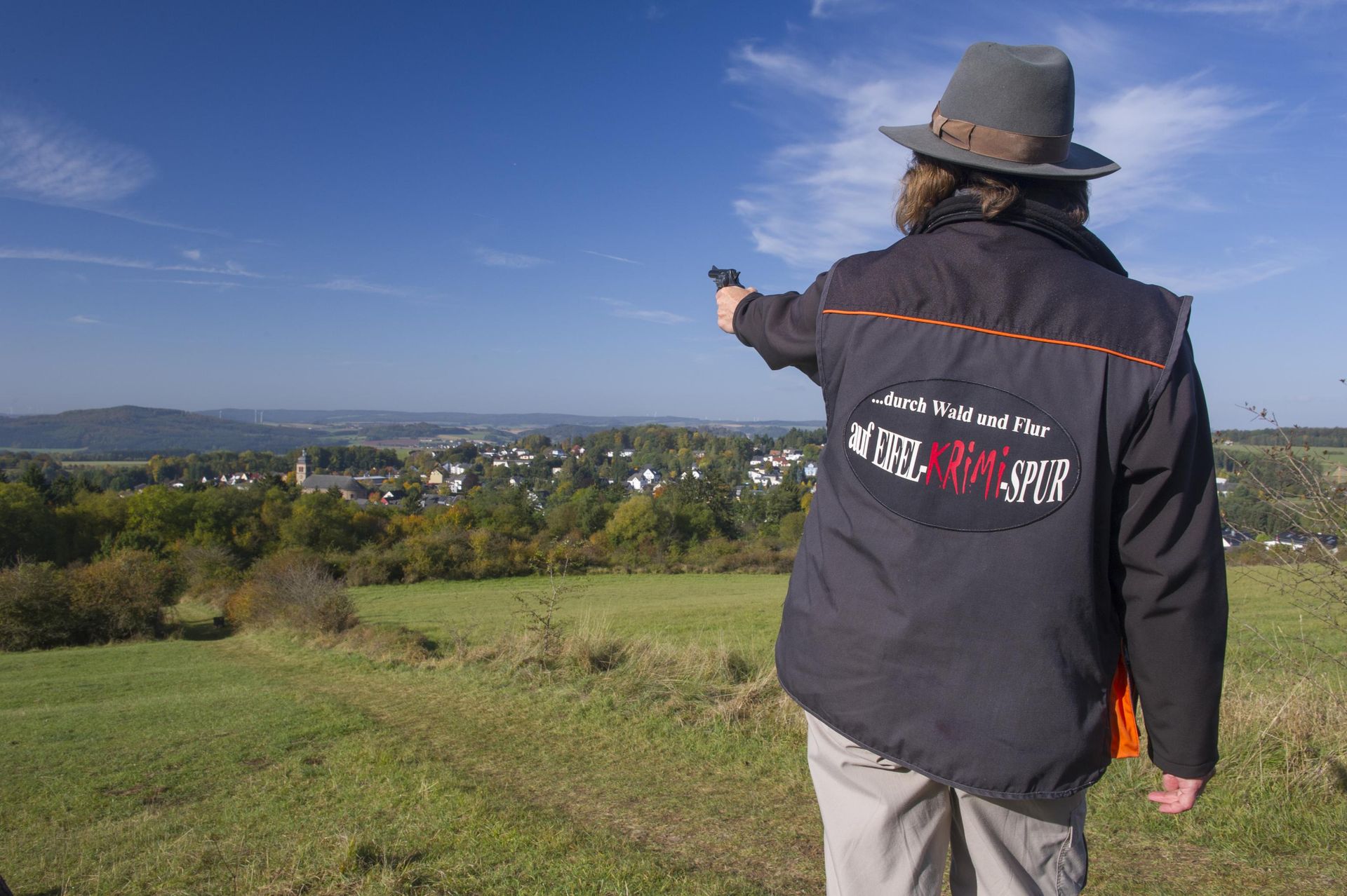 Eine Hut tragende Person steht mit dem Rücken zum Betrachter und zeigt mit einer Pistole in die Ferne. Sie trägt eine Jacke mit der Aufschrift "...durch Wald und Flur auf Eifel-Krimi-Spur" auf dem Rücken. Vor der Person liegt eine große Wiese und dahinter in der Ferne ein Dorf mit  vielen Häusern.