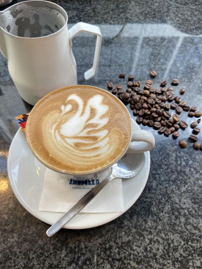 Eine Tasse Cappuccino mit Latte Art auf einem Tisch, daneben Kaffeebohnen und ein Milchkännchen.