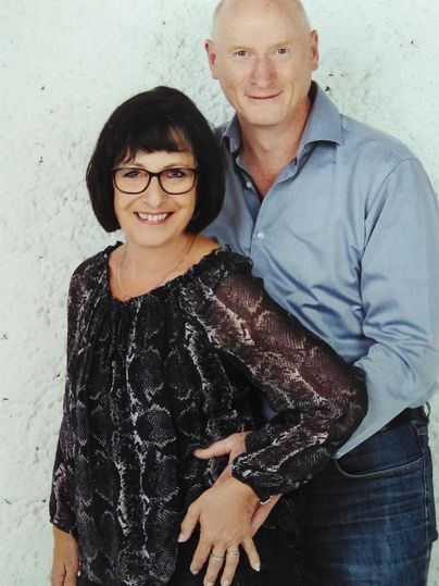 Ein Paar posiert lächelnd vor einer neutralen Wand. Die Frau trägt eine schwarze Bluse, der Mann ein hellblaues Hemd.