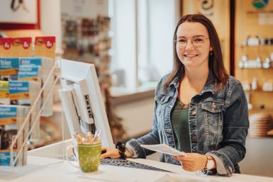 Eine Frau steht an einem hohen Schreibtisch mit Computer und Tastatur vor sich und lächelt in die Kamera. Hinter ihr ist der Souveniershop und einige Prospektregale der Tourist-Information.