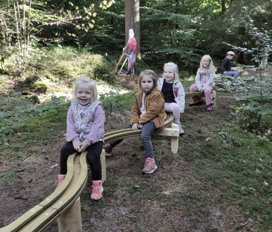 Fünf Kinder sitzen auf der Holzkugelbahn im Wald. Sie lachen und genießen die Natur. Im Hintergrund ist eine Figur eines Waldarbeiters zu sehen., © Touristik GmbH Gerolsteiner Land