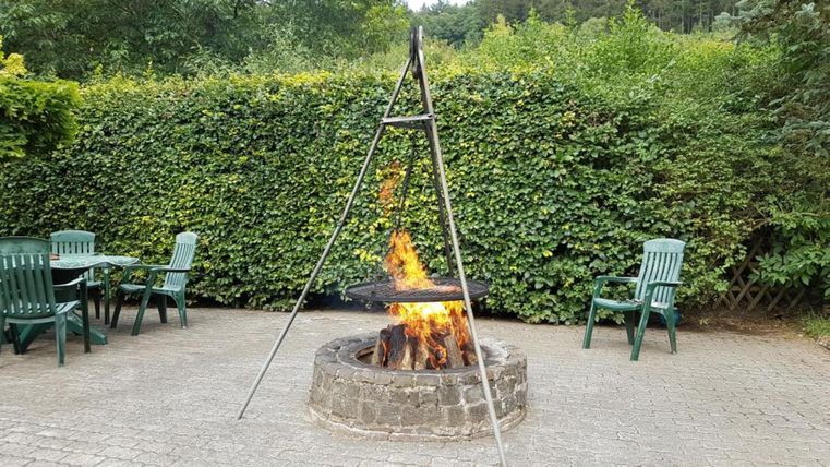 Ein gemütliches Lagerfeuer in einem Steinkreis steht unter einem dreibeinigen Gestell. Im Hintergrund sind grüne Pflanzen und einige Sitzgelegenheiten aus Plastik zu sehen.