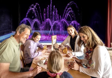 A man and a woman are sitting at a table with their three daughters, drinking coffee or cold drinks. Behind the family, a colourful water feature organ sprays water into the air.