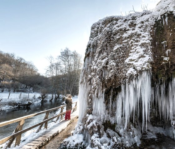 Gefrorener Nohner Wasserfall mit Eiszapfen, eine Person auf einem Steg, winterliche Landschaft mit Schnee und kahlen B&auml;umen