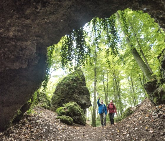 Two hikers walk through a forest, photographed from a cave. The surroundings are green and wooded., © Eifel Tourismus GmbH, Dominik Ketz