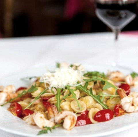 Auf einem Teller angerichtetes Nudelgericht mit Cherrytomaten, Garnelen, Rucola und Parmesan. Daneben auf einer Serviette liegendes Besteck und ein Glas Rotwein.