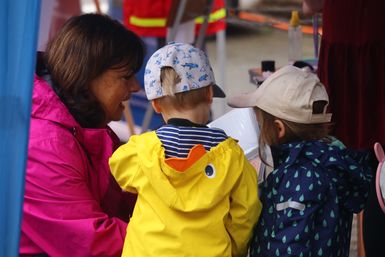 Zwei Kinder in Regenjacken stehen neben einer Frau, die neben ihnen hockt und schauen sich etwas an.