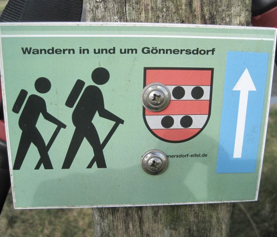 Green hiking sign with two stylized hikers, a coat of arms and a blue arrow. Text: "Hiking in and around Gönnersdorf"., © Touristik GmbH Gerolsteiner Land, Ute Klinkhammer