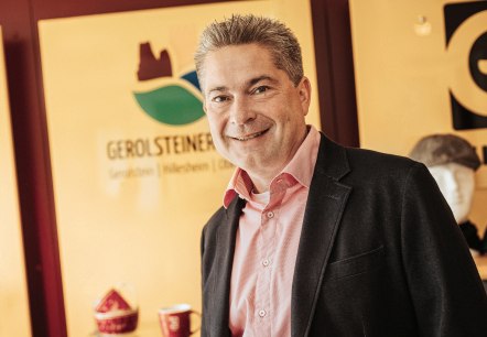 Ein schick gekleideter Herr mit Hemd und Sakko l&auml;chelt in die Kamera. Hinter ihm das Logo der Touristik GmbH Gerolsteiner Land und einige Souvenierartikel.