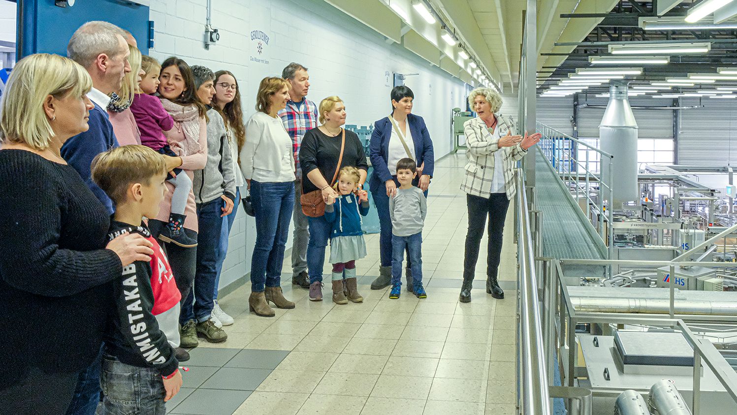 Eine Personengruppe steht auf einer Empore in einer Produktionshalle und hört einer Damen zu.