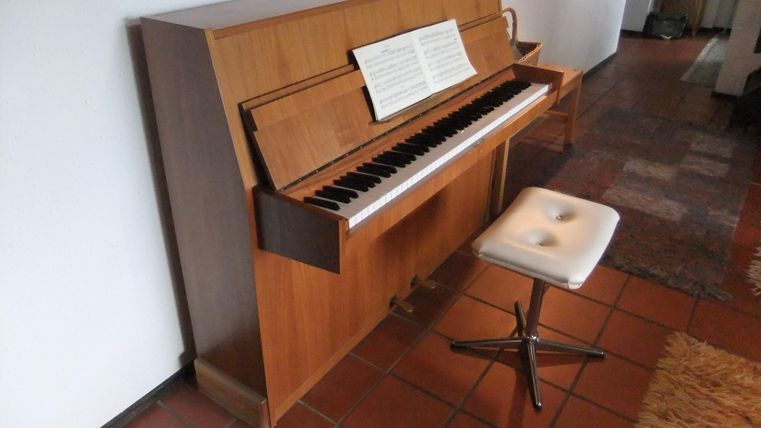 Ein Klavier aus Holz steht in einem Raum. Daneben befindet sich ein Hocker mit einem weißen Sitzpolster.