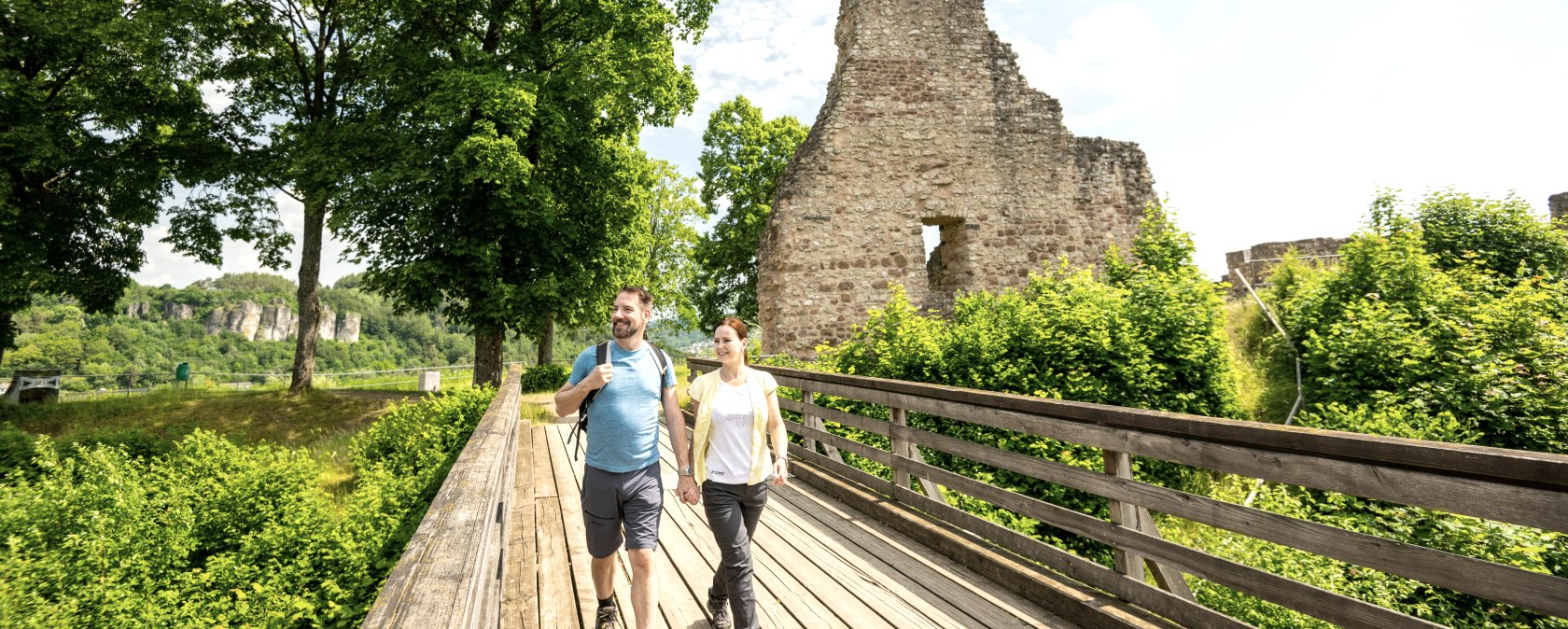 Een gedeeltelijk bewaard gebleven kasteelru&iuml;ne omringd door bloeiende bomen en struiken. Een houten loopbrug met twee wandelaars erop leidt naar het kasteel.